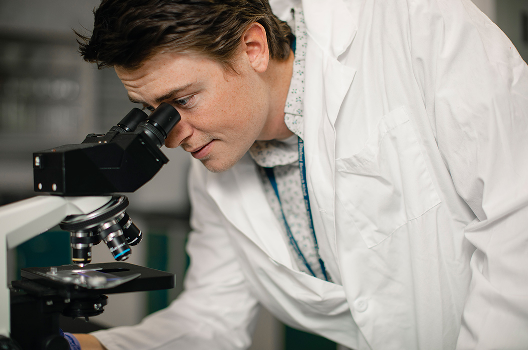 biomedical student in white lab coat leaning down looking into a microscope