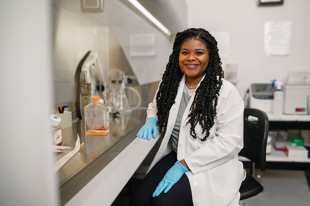 biomedical student sitting in a lab with rubber gloves on infront of lab equipment smiling in a white lab coat