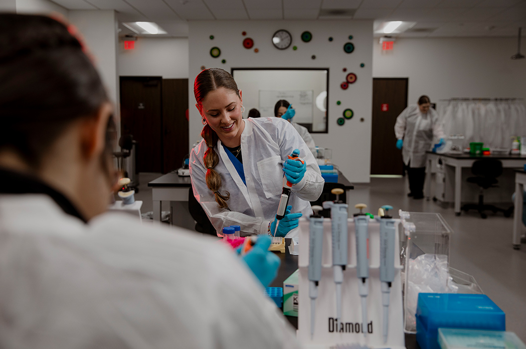 health professions student in white coat using syringe equipment standing in a lab room