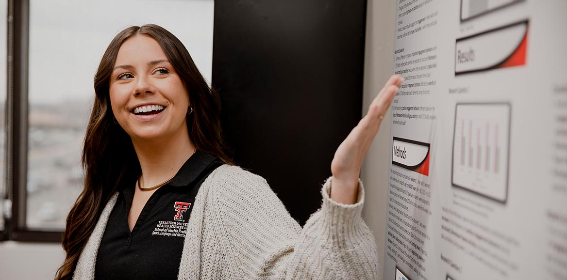 TTUHSC student standing infront of a presentation board with her hand pointing towards it smiling