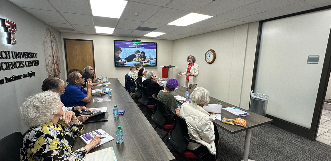 group of people sitting at a desk looking toward the front at the presenter that is standing infront of a board