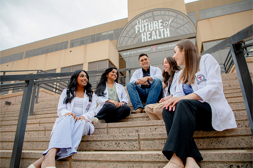 Five medical students wearing white coats sit together on outdoor stairs in front of the Texas Tech University Health Sciences Center building.