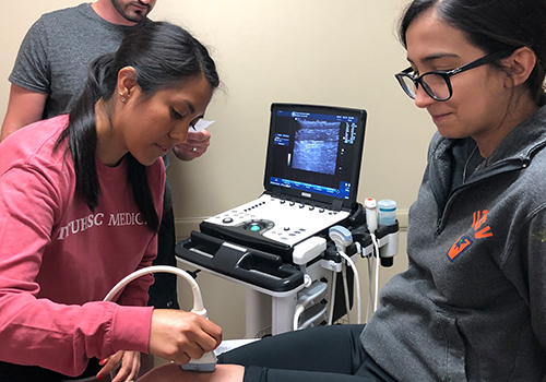 TTUHSC students in a patient room one sitting on a table while the other holds a transducer on the knee of the sitting student TTUHSC students in a patient room one sitting on a table while the other holds a transducer on the knee of the sitting student