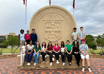 A group of TTUHSC Summer Enrichment students sitting on concrete wall infront of TTUHSC seal