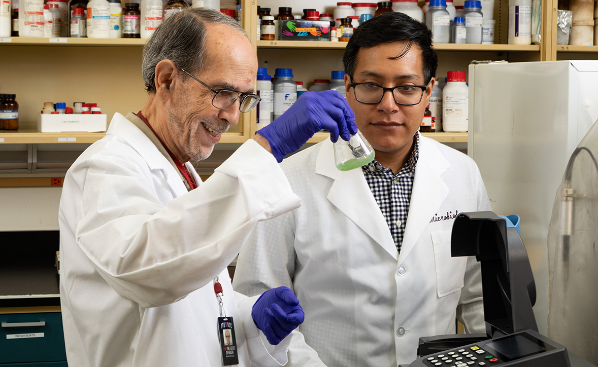 Two scientists in lab coats work together in a laboratory. One is holding a flask containing a green liquid, smiling as he examines it, while the other observes attentively. Shelves lined with various bottles and lab supplies are visible in the background.