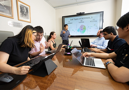 seven students in a dry lab room on laptops sitting around a table watching a presenter standing by a screen seven students in a dry lab room on laptops sitting around a table watching a presenter standing by a screen