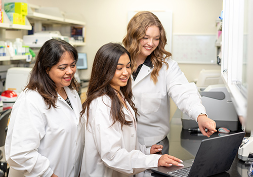 three women in white lab coats smiling and staring at laptop screen in a lab three women in white lab coats smiling and staring at laptop screen in a lab