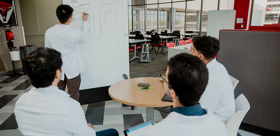 four TTUHSC students in white lab coats sitting at a table while one student stands and writes on a dry erase marker