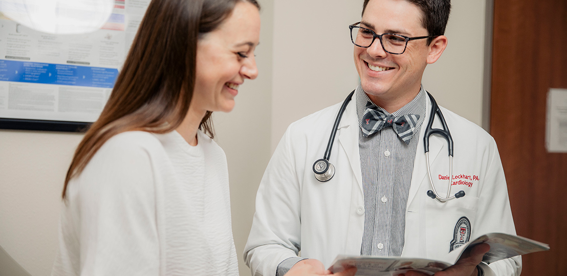 TTUHSC faculty member in white lab coat standing, smiling infront of patient sitting on a patient table in a grey tshirt holding a book open while the patient looks at it