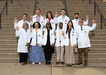 TTUHSC medical education students standing in white lab coats infront of the Lubbock campus stairway outdoors with the guns up made by their hands