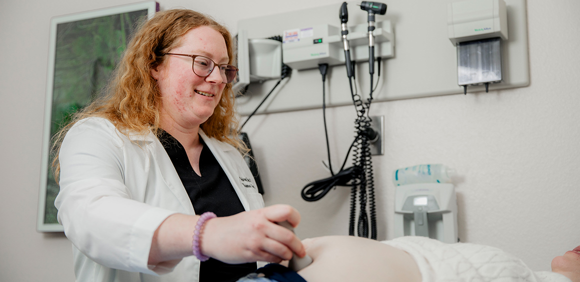TTUHSC student in white lab coat holding ultrasound machine on pregnant patients stomach who is lying on a pateint table in a patient room