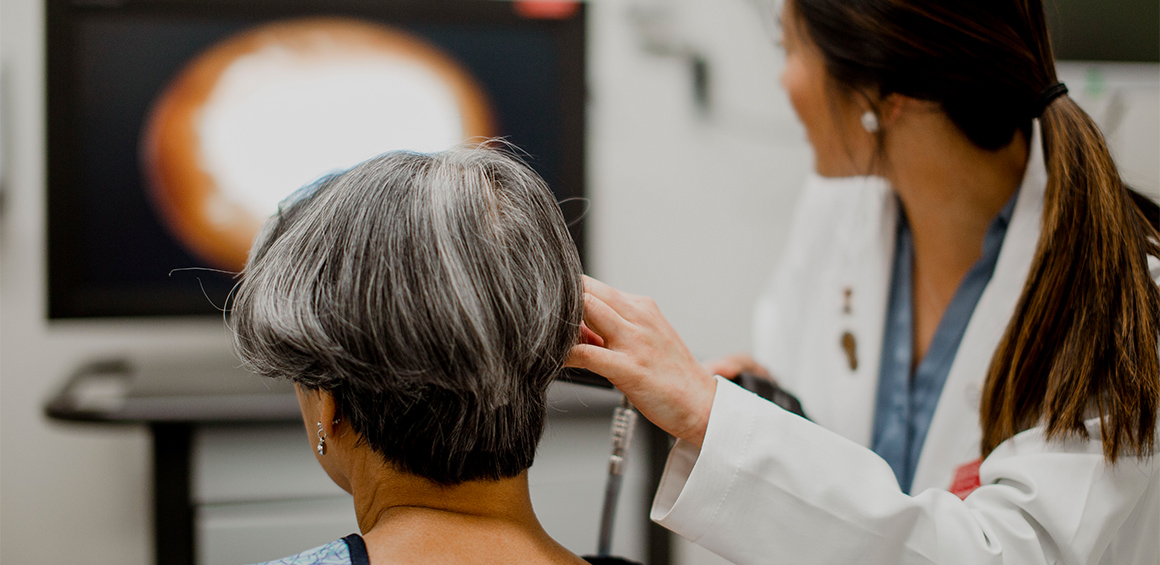 TTUHSC student in white lab coat standing over a patient in a bed holding an otoscope on the patients ear.