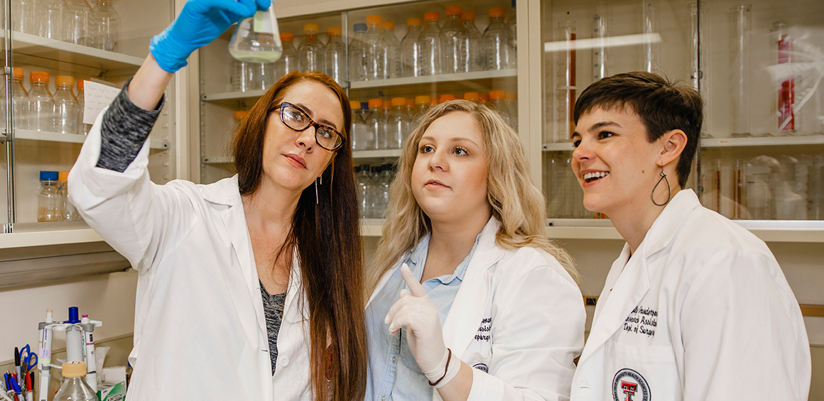 TTUHSC BCoRE Director Kendra Rumbaugh holding a glass jar in the air while two TTUHSC students look up at it standing next to her
