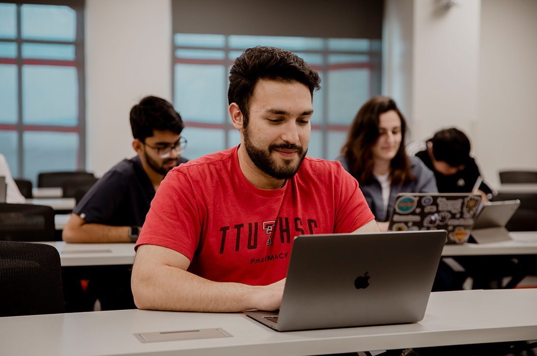 pharmacy student sitting at a desk, working on a computer in a classroom with other students seated behind at their desks