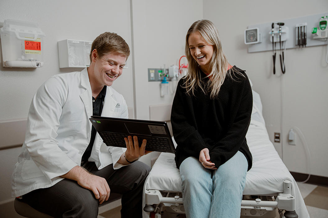 pharmacy student in white labcoat holding computer showing patient sitting on hospital bed in patient room smiling